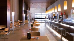 La moderna cafetería del Hotel Silken Puerta de Valencia cuenta con muebles de madera, una larga barra, lámparas colgantes y ventanas llenas de luz, creando un escape acogedor para los visitantes cerca de la Puerta de Valencia. - ForTwo - Planes para parejas