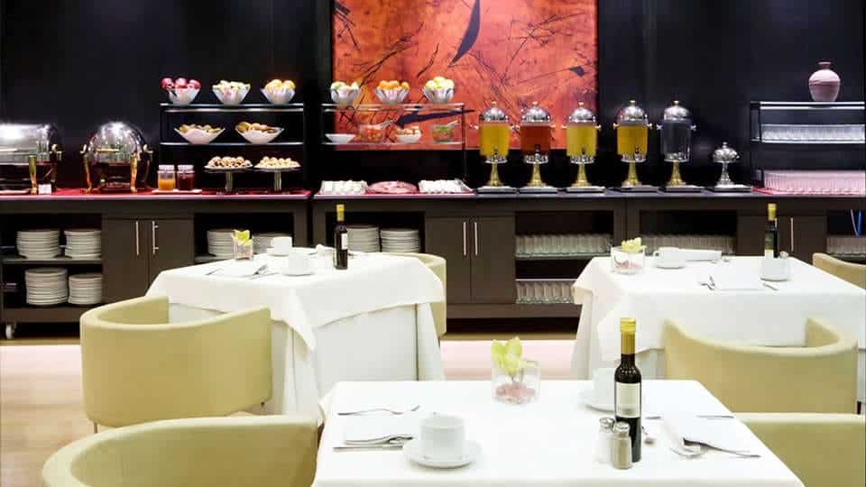 La Escapada con desayuno, spa, cava y frutas en el Hotel H10 Marina 4* de Barcelona ofrece una experiencia culinaria ideal con mesas elegantemente dispuestas, botellas de vino y un suntuoso buffet con diversos platos y dispensadores de zumos después de un relajante día de spa. - ForTwo - Planes para parejas