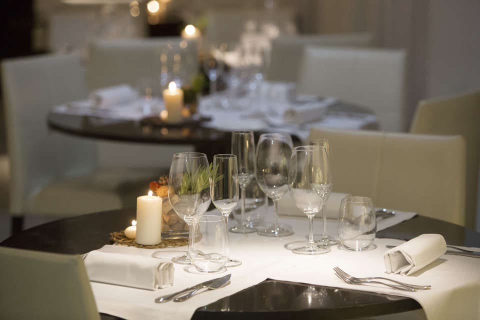 La Escapada Hospes Palau de la Mar con desayuno y spa (Valencia) ofrece un ambiente elegante con velas, cristalería, servilletas blancas y cubiertos en mesas redondas en una sala suavemente iluminada, perfecta para una experiencia íntima. - ForTwo - Planes para parejas
