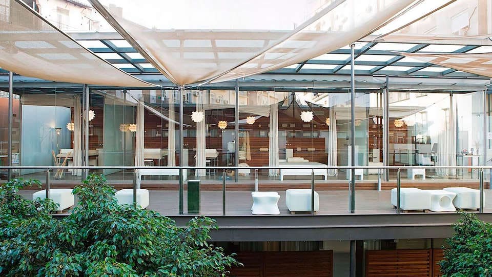 El moderno edificio de cristal, con amplios ventanales y elegantes marquesinas, se alza elegantemente entre exuberantes plantas, haciéndose eco de la estética tranquila de Escapada Hospes Palau de la Mar con desayuno y spa (Valencia). - ForTwo - Planes para parejas