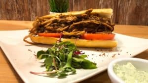 Disfrute de un sándwich de cerdo desmenuzado con tomate y verduras en un plato blanco, acompañado de salsa verde en el Restaurante Galipán en Valencia. - ForTwo - Planes para parejas
