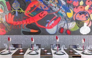 En el Hotel Silken Puerta de Valencia, un vibrante mural de mariscos adorna la pared sobre una mesa de comedor, realzado por centros de mesa florales rojos y blancos. - ForTwo - Planes para parejas