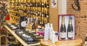 El elegante interior de la vinoteca presenta botellas elegantemente dispuestas en mesas y estantes, con un set de champán en caja como protagonista. El encanto de la "Cata de vermuts con aperitivo en Di-Ví (Barcelona)" invita a explorar la selección de vermuts, ideal para una experiencia de aperitivo perfecta. - ForTwo - Planes para parejas
