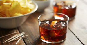 La Cata de vermuts con aperitivo en Di-Ví en Barcelona queda perfectamente capturada con una copa de cóctel de vermut oscuro con hielo y aceitunas junto a un bol de patatas fritas y palillos, ideal para un delicioso momento de aperitivo. - ForTwo - Planes para parejas