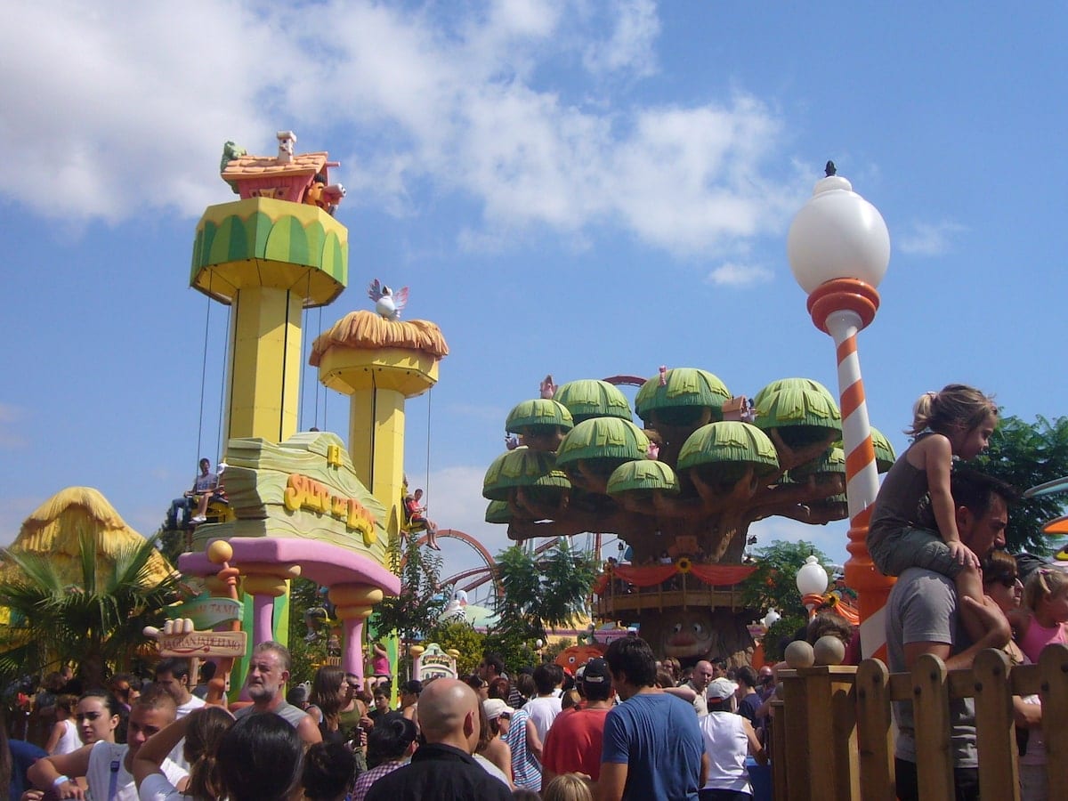 El animado parque de atracciones, similar al Caribe Aquatic Park de Port Aventura en Tarragona, presenta vibrantes atracciones con forma de árbol y multitudes enérgicas bajo un cielo azul brillante. - ForTwo - Planes para parejas