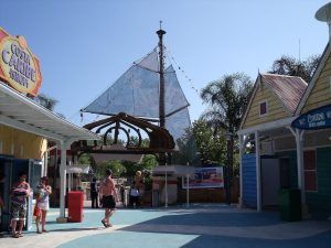 La zona del parque de atracciones al aire libre, fiel al ambiente de Tarragona, luce una decoración con mástiles de veleros. Los visitantes disfrutan del animado ambiente y observan los carteles de las tiendas cercanas, que evocan un día con "Entradas para el parque acuático Caribe Aquatic Park de Port Aventura (Tarragona)". - ForTwo - Planes para parejas
