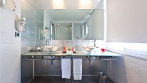 Disfrute de un baño elegante en Escapada OD Ocean Drive con desayuno (Ibiza), con dos lavabos, un gran espejo y una encimera de cristal. Disfrute de la comodidad de un secador de pelo de pared y una iluminación brillante para un ambiente elegante. - ForTwo - Planes para parejas