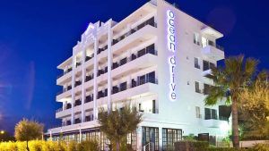 Este hotel blanco de varios pisos con balcones y un letrero de neón de OD Ocean Drive captura el vibrante ambiente ibicenco del Escapada OD Ocean Drive con desayuno (Ibiza), realzado por una palmera que se mece suavemente. - ForTwo - Planes para parejas