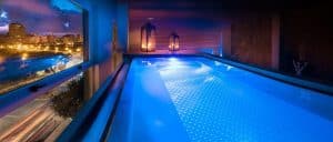 Disfrute de un baño nocturno en la piscina cubierta del Hotel SH Valencia Palace, iluminada con luces azules y velas. Relájese con la impresionante vista de la ciudad a través del amplio ventanal al atardecer: la escapada perfecta en nuestro hotel de Valencia. - ForTwo - Planes para parejas
