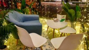 Un acogedor rincón de cafetería con un sillón azul, dos sillas blancas y una mesa con plantas en macetas y luces de cadena evoca el encanto de Ibiza, haciéndolo sentir como una joya escondida del Restaurante Ibiza Poke. - ForTwo - Planes para parejas