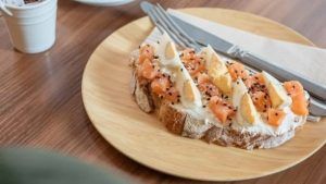 Un sándwich abierto con queso crema, salmón ahumado, huevos cocidos y semillas de sésamo en un plato de madera ofrece una delicia al estilo ibicenco que encaja perfectamente con el ambiente moderno del Restaurante Ibiza Poke. - ForTwo - Planes para parejas