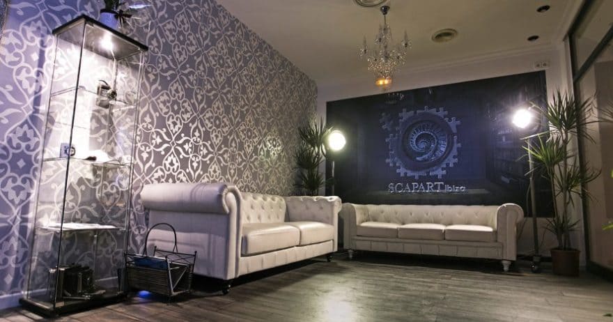 Elegante habitación con dos sofás blancos, papel pintado estampado, una vitrina de cristal y una lámpara de araña, evocando el ambiente de un Scapart - Escape Room en Ibiza. - ForTwo - Planes para parejas