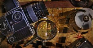 Una cámara vintage, una lupa y rollos de película yacen sobre un viejo mapa de Ibiza, evocando aventuras pasadas. La escena evoca el comienzo de un desafío de Scapart - Escape Room (Ibiza), con una correa de cuero marrón que realza el misterio. - ForTwo - Planes para parejas