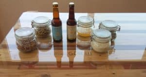 Tarros de grano y dos botellas de cerveza descansan sobre una mesa de cristal con marco de madera, capturando la esencia viva de Valencia e invitándote a unirte a la experiencia de Visita a la fábrica de cerveza Gènesis con cata y almuerzo. - ForTwo - Planes para parejas