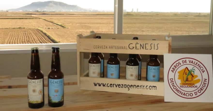 Las botellas de cerveza artesanal se encuentran en una caja de madera con la etiqueta Gènesis junto a una ventana con vista al campo de Valencia, invitándolo a la "Visita a la fábrica de cerveza Gènesis con cata y almuerzo (Valencia)" para una experiencia de degustación exquisita. - ForTwo - Planes para parejas