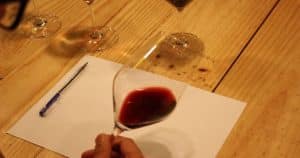 En Barcelona, una persona sostiene una copa de vino tinto sobre un papel en blanco sobre una mesa de madera, rodeada de bolígrafos y más vasos, preparando el escenario para una íntima "Cata de vinos en Barcelona". - ForTwo - Planes para parejas
