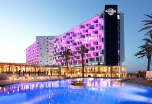 El Hard Rock Hotel (Ibiza) cuenta con una iluminación rosa brillante que se refleja en una espaciosa piscina, rodeada de palmeras al anochecer. - ForTwo - Planes para parejas