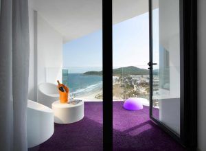 El balcón contemporáneo del Hard Rock Hotel de Ibiza ofrece una vista al mar, resaltada por sillas blancas, una alfombra violeta y una mesa con una botella de champán en una hielera naranja. - ForTwo - Planes para parejas