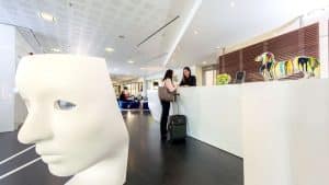 En el vibrante vestíbulo del Hotel Náutico Ebeso en Ibiza, una escultura abstracta de un rostro blanco se encuentra junto a una colorida estatua de un perro mientras los huéspedes se registran ansiosos. - ForTwo - Planes para parejas