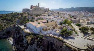 Una vista aérea de Ibiza muestra una ciudad costera con edificios históricos sobre un acantilado, rodeada de vegetación y con vistas al mar, cerca del famoso Hotel Mirador de Dalt Vila. - ForTwo - Planes para parejas