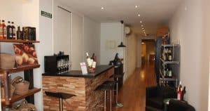 La Cata de vinos en Barcelona ofrece un acogedor bar con un mostrador de madera, taburetes y botellas de vino alineadas en los estantes en un pasillo estrecho y bien iluminado, capturando el encanto de las joyas ocultas de Barcelona para una cata de vinos íntima. - ForTwo - Planes para parejas
