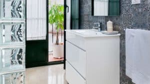 Un baño moderno luce un tocador blanco, azulejos decorativos y una elegante pared de cristal. Una maceta cerca de la puerta añade un toque natural, similar a una escapada con desayuno y crucero en barco por el Guadalquivir. - ForTwo - Planes para parejas