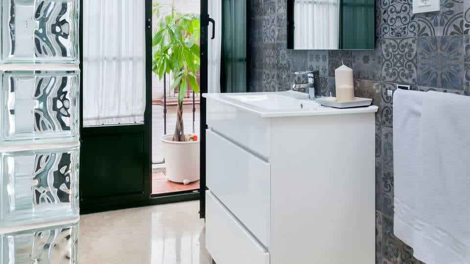 Un baño moderno luce un tocador blanco, azulejos decorativos y una elegante pared de cristal. Una maceta cerca de la puerta añade un toque natural, similar a una escapada con desayuno y crucero en barco por el Guadalquivir. - ForTwo - Planes para parejas