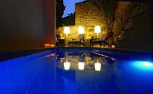 La piscina iluminada del Hotel Mirador de Dalt Vila en Ibiza, con sillas a su alrededor y lámparas brillantes sobre un fondo de pared de piedra, ofrece un refugio tranquilo por la noche. - ForTwo - Planes para parejas