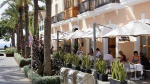 Una cafetería al aire libre en Ibiza ofrece comensales bajo sombrillas, una fachada de color melocotón similar a la del Hotel Mirador de Dalt Vila y palmeras que bordean el paseo. - ForTwo - Planes para parejas