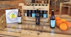 Una caja de botellas de cerveza Gènesis descansa sobre una mesa de madera, rodeada de jugosas naranjas y tallos de arroz frescos, personificando la esencia perfecta de una "Visita a la fábrica de cerveza Gènesis con cata y almuerzo" en Valencia. - ForTwo - Planes para parejas