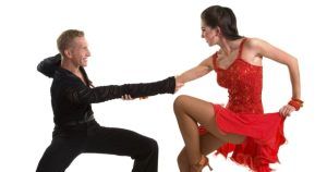 Un hombre y una mujer con atuendo de baile posan con energía, tomados de la mano, como si estuvieran en medio de una animada rutina de bachata. La mujer deslumbra con un vibrante vestido rojo, capturando el espíritu de las noches madrileñas, similar a "Cuatro clases de salsa o bachata nivel iniciación (Madrid)". - ForTwo - Planes para parejas