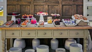 Una mesa de madera repleta de pasteles, pan y postres se encuentra bellamente colocada contra una pared rústica, capturando la esencia de un hotel Escapada H10 Montcada con extras (Barcelona). Perfecto para saborear estas delicias. - ForTwo - Planes para parejas