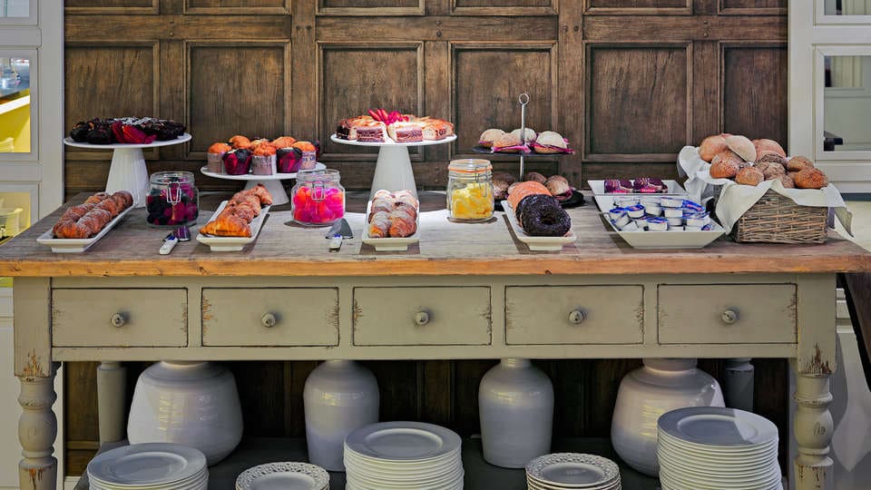 Una mesa de madera repleta de pasteles, pan y postres se encuentra bellamente colocada contra una pared rústica, capturando la esencia de un hotel Escapada H10 Montcada con extras (Barcelona). Perfecto para saborear estas delicias. - ForTwo - Planes para parejas