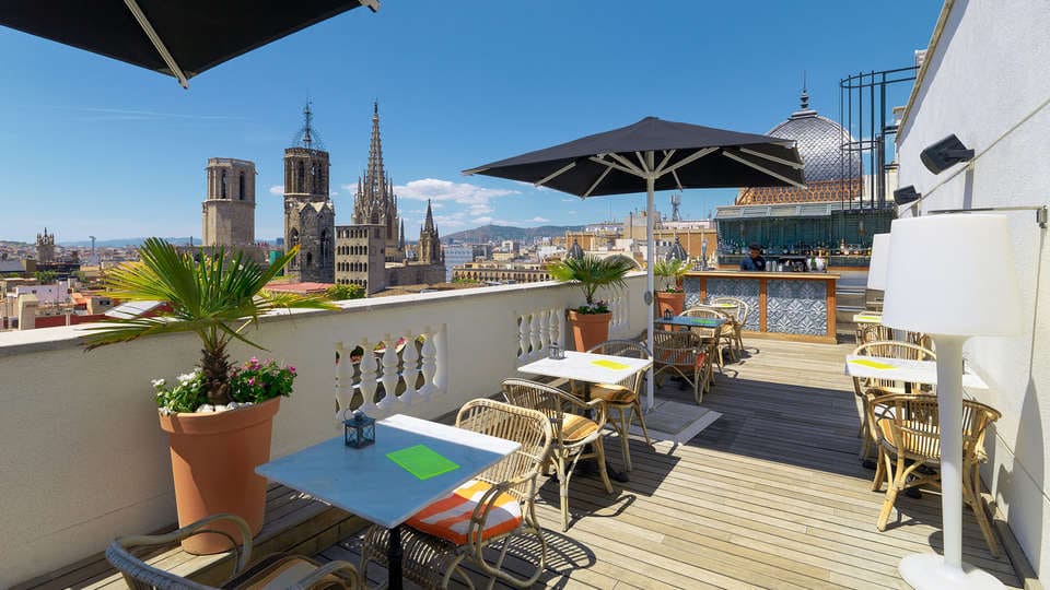 La terraza de la azotea del hotel Escapada H10 Montcada en Barcelona ofrece mesas, sillas y sombrillas con impresionantes vistas a torres históricas contra un cielo azul claro, ideal para su escapada. - ForTwo - Planes para parejas