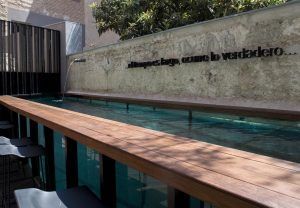 Disfrute del encanto del Hotel Caro (Valencia) con modernos asientos al aire libre, un bar de madera junto a una piscina estrecha, una tranquila fuente de pared y la exuberante sombra de los árboles. - ForTwo - Planes para parejas