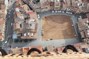 Vista desde un Vuelo estándar en globo por Cataluña con Globus (Barcelona), la perspectiva aérea muestra una plaza de Cataluña rodeada de edificios, con una zona central rústica de tierra salpicada de coches estacionados. - ForTwo - Planes para parejas