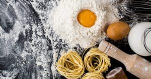Sobre una superficie oscura reposan harina, huevo y masa de pasta; cerca hay un rodillo, un batidor y cáscaras de huevo, capturando el arte del Curso de pasta fresca en Madrid con degustación. - ForTwo - Planes para parejas