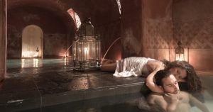 Dos personas se relajan junto a una piscina cubierta poco iluminada, evocando el ambiente del "Hammam en Córdoba: baño árabe con kessa y masaje MIDRA 30", con una gran linterna decorativa que realza la atmósfera serena. - ForTwo - Planes para parejas