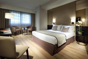 Disfrute de la comodidad de una habitación moderna en el Hotel Eurostars Rey Don Jaime (Valencia), equipada con una cama doble, dos sillas, una mesa y una ventana con cortinas transparentes. La suave iluminación aporta calidez y crea un ambiente acogedor. - ForTwo - Planes para parejas
