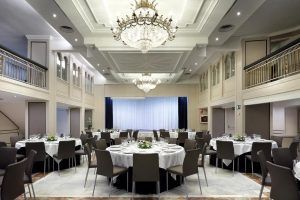 El elegante salón de banquetes del Hotel Eurostars Rey Don Jaime en Valencia luce mesas redondas con manteles blancos, exquisitas lámparas de araña y un escenario adornado con cortinas. - ForTwo - Planes para parejas