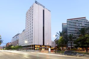 Un edificio hotelero alto y moderno con palmeras y una calle animada en primer plano bajo el cielo despejado de Valencia, que recuerda el ambiente lujoso del Hotel Eurostars Rey Don Jaime. - ForTwo - Planes para parejas