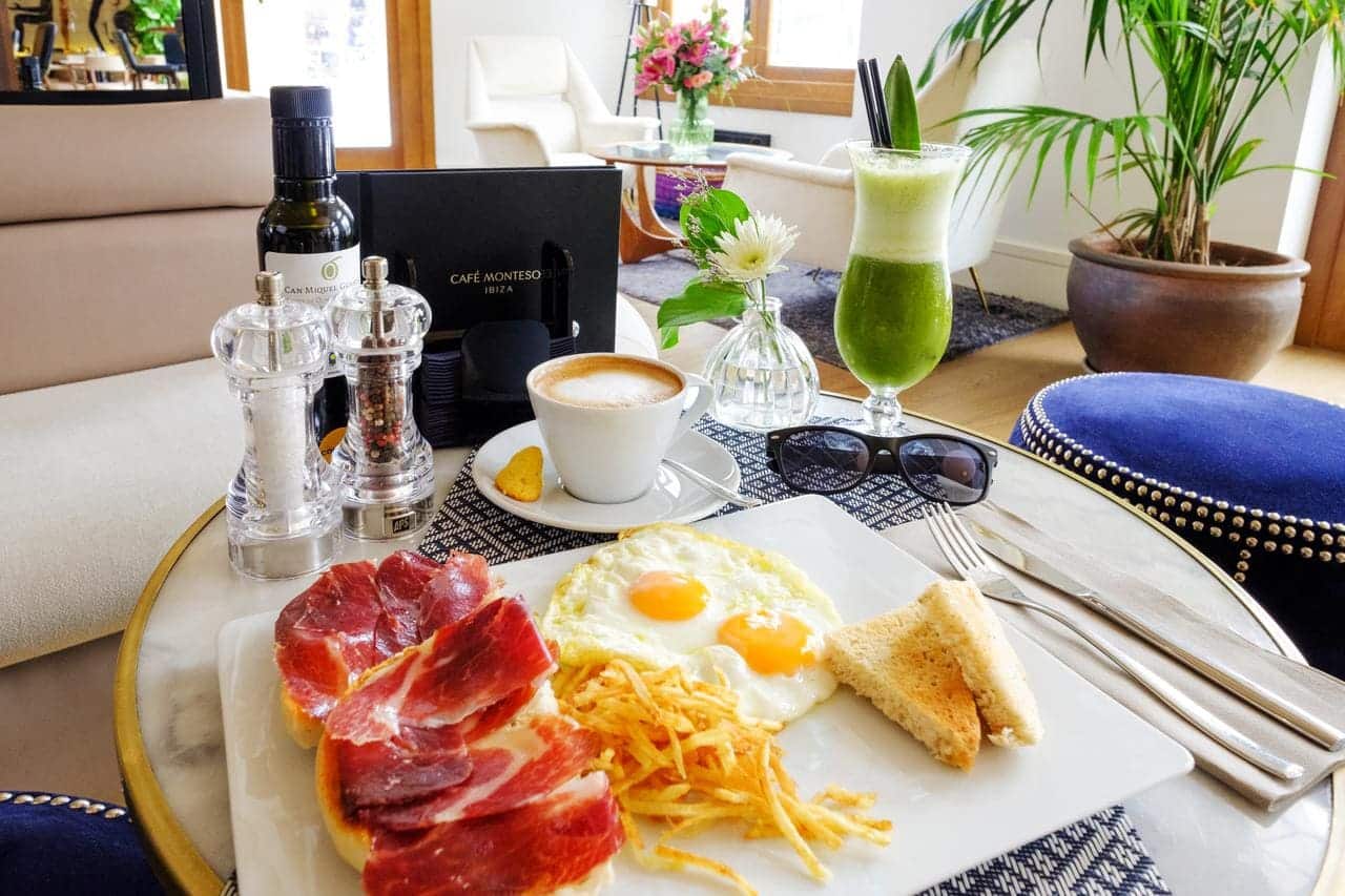 En el Gran Hotel Montesol: Curio Collection by Hilton de Ibiza, se sirve un elegante desayuno con huevos, jamón, tostadas, patatas fritas, café y zumo verde. Las gafas de sol descansan sobre la mesa mientras la luz del sol inunda este vibrante espacio. - ForTwo - Planes para parejas