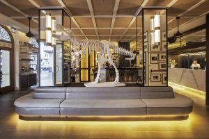 El moderno vestíbulo del Gran Hotel Montesol: Curio Collection by Hilton en Ibiza cuenta con un elegante esqueleto de dinosaurio exhibido en un sofá curvo e iluminado, junto a un elegante mostrador de recepción de mármol. - ForTwo - Planes para parejas