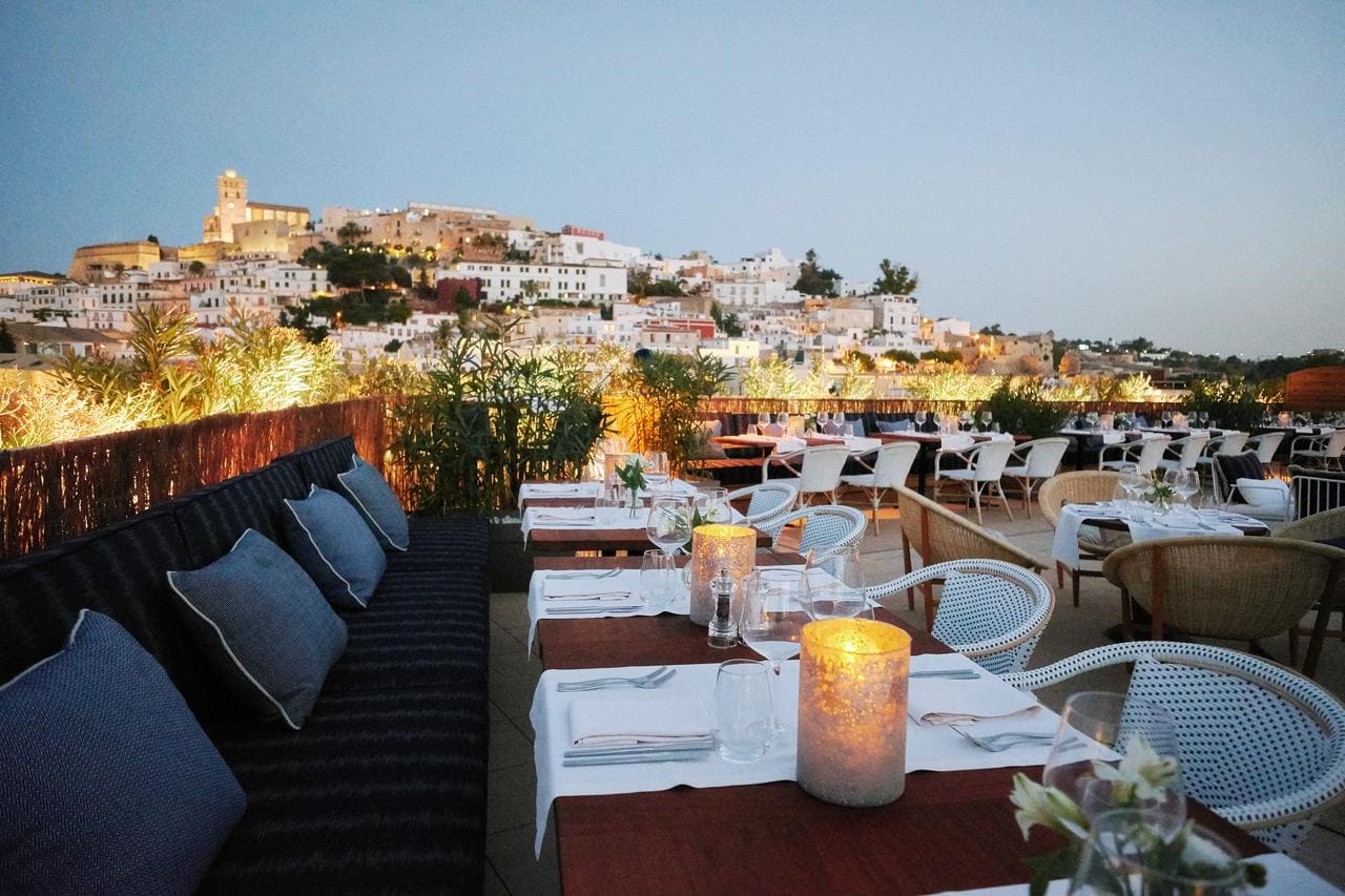 Descubra el mágico ambiente del restaurante en la azotea del Gran Hotel Montesol: Curio Collection by Hilton (Ibiza). Disfrute de las impresionantes vistas de la ciudad al atardecer, rodeado de elegantes mesas blancas y cómodos asientos, creando un oasis de sofisticación en esta joya escondida. - ForTwo - Planes para parejas
