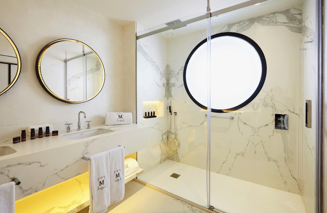 Experimente la elegancia de un baño moderno de inspiración ibicenca con espejos redondos, encimeras de mármol y una ducha de cristal con una gran ventana circular. Disfrute del lujo que evoca al Gran Hotel Montesol: Curio Collection by Hilton en Ibiza. - ForTwo - Planes para parejas