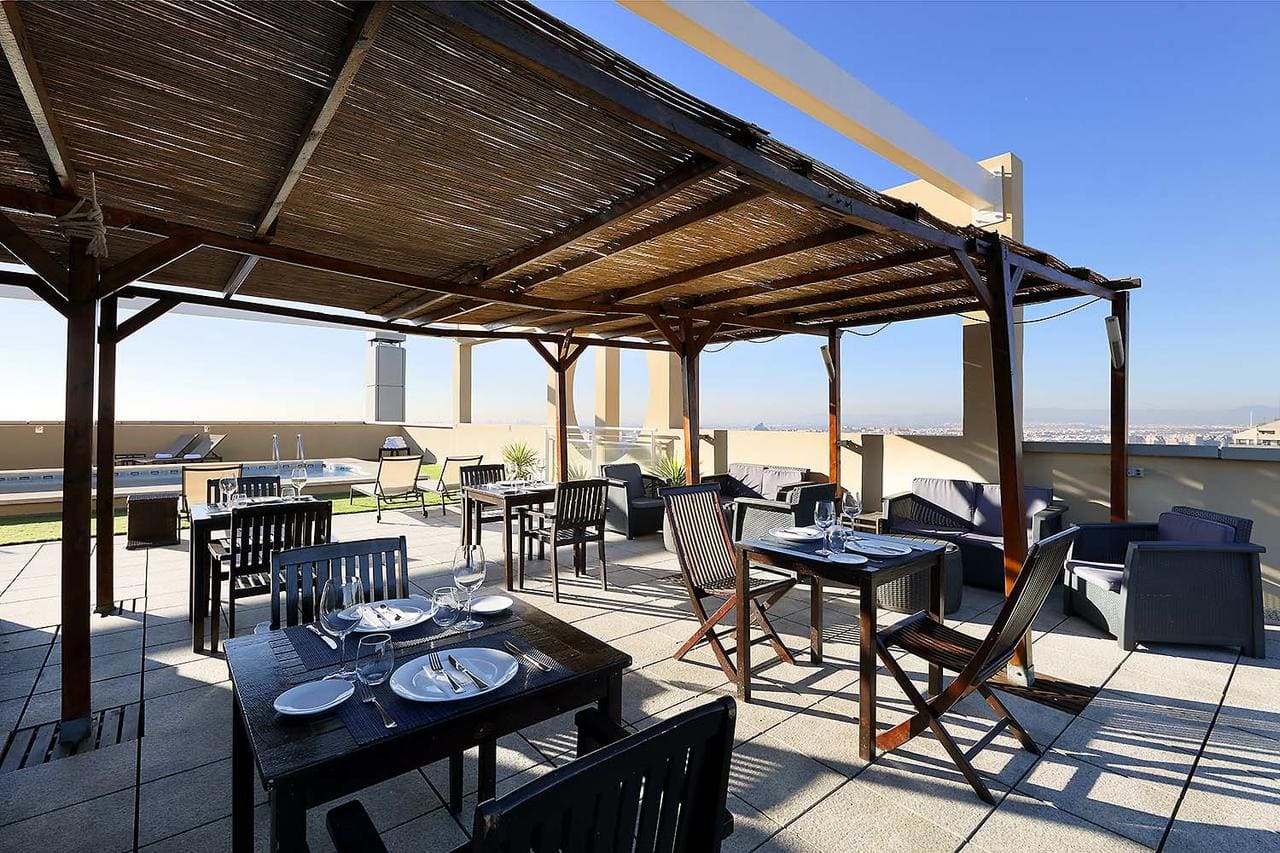 El Hotel Eurostars Gran Valencia cuenta con un hermoso patio en la azotea con mesas y sillas de comedor de madera bajo una pérgola, que ofrece vistas al cielo despejado. - ForTwo - Planes para parejas