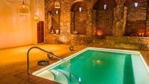Una piscina cubierta con agua turquesa brillante, paredes de ladrillo e iluminación ambiental de lámparas tejidas y velas reproduce el ambiente sereno de Escapada de 2 noches + cena romántica + Hammam (Córdoba). - ForTwo - Planes para parejas