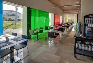 Una moderna cafetería cuenta con paneles de vidrio de colores, mesas dispuestas y una estación de café, todo con vista al césped verde y al impresionante Hotel Barceló Valencia. - ForTwo - Planes para parejas