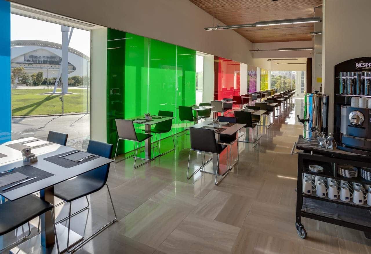 Una moderna cafetería cuenta con paneles de vidrio de colores, mesas dispuestas y una estación de café, todo con vista al césped verde y al impresionante Hotel Barceló Valencia. - ForTwo - Planes para parejas