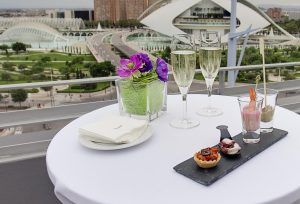 Desde el elegante Hotel Barceló de Valencia, una mesa preparada con copas de champán, aperitivos y flores violetas ofrece una impresionante vista del futurista horizonte de la ciudad. - ForTwo - Planes para parejas